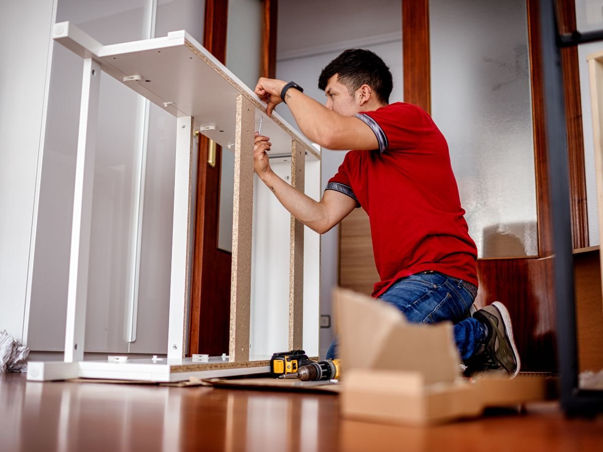 A man is focused on assembling a wooden piece of furniture in a well-lit workshop. Tools are scattered around him.