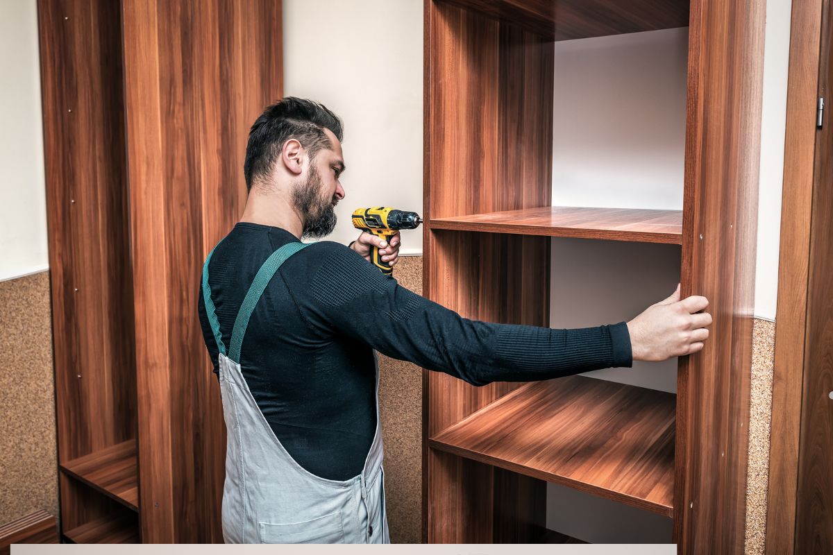 A person in overalls uses a power drill to assemble a wooden bookshelf against a light-colored wall.