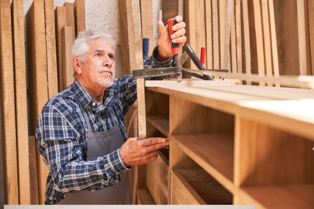 An elderly man in a plaid shirt works on woodworking, measuring and adjusting parts of a wooden shelf in a workshop.